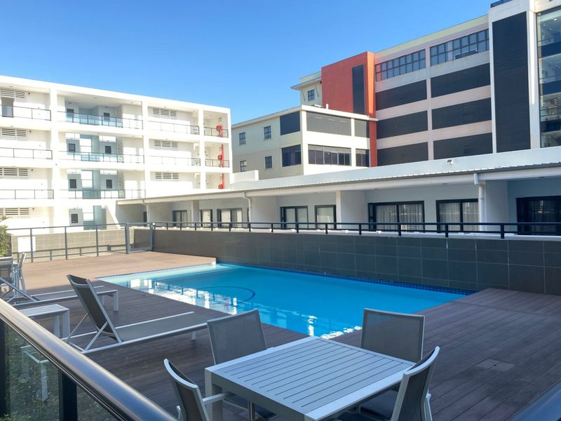 A communal swimming pool area featuring a timber deck, modern tiled retaining wall, and surrounding apartment building views.