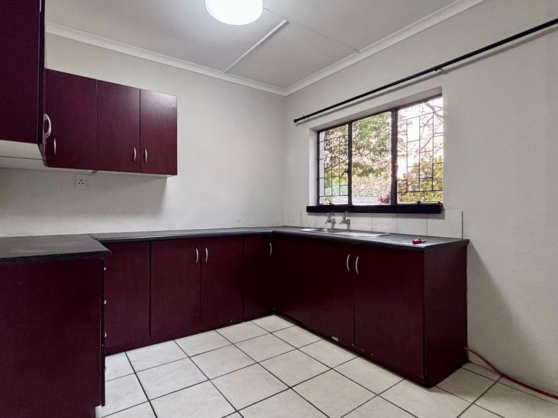 A functional kitchen featuring dark laminate cabinetry, contrasting countertops, tiled flooring, and a window with security bars.