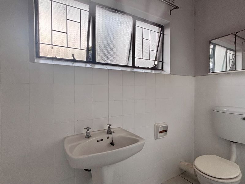 A functional bathroom featuring a pedestal basin, toilet, and shower, with white wall tiling and a window providing natural light.