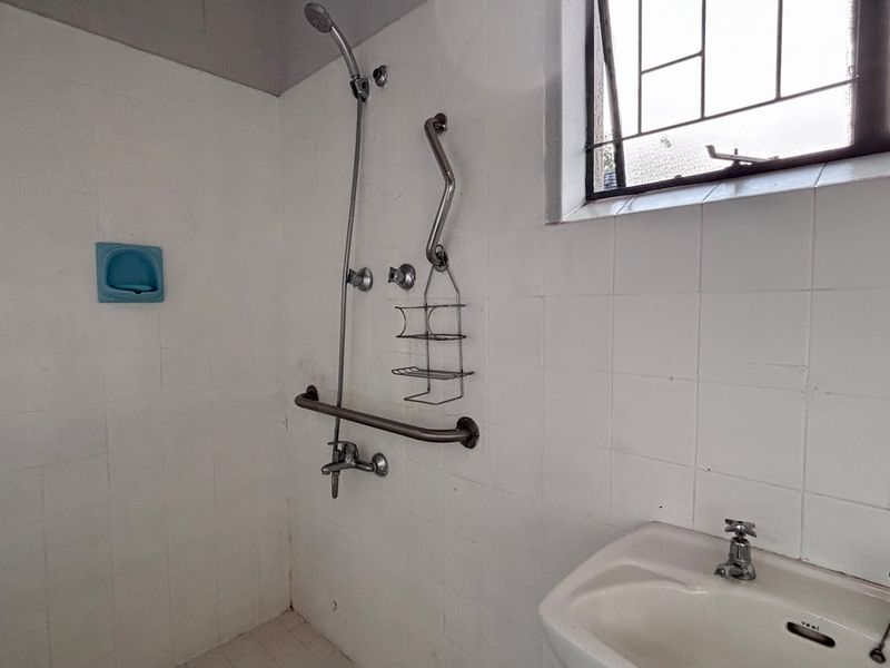 A functional bathroom featuring a shower with grab rails, a wall-mounted basin, and tiled walls, requiring cosmetic updates to improve its overall condition.