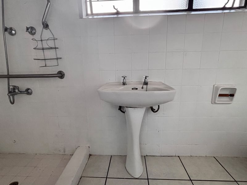 A functional bathroom featuring a pedestal basin, shower area with grab rail, and tiled finishes, requiring cosmetic updates to improve its overall condition.