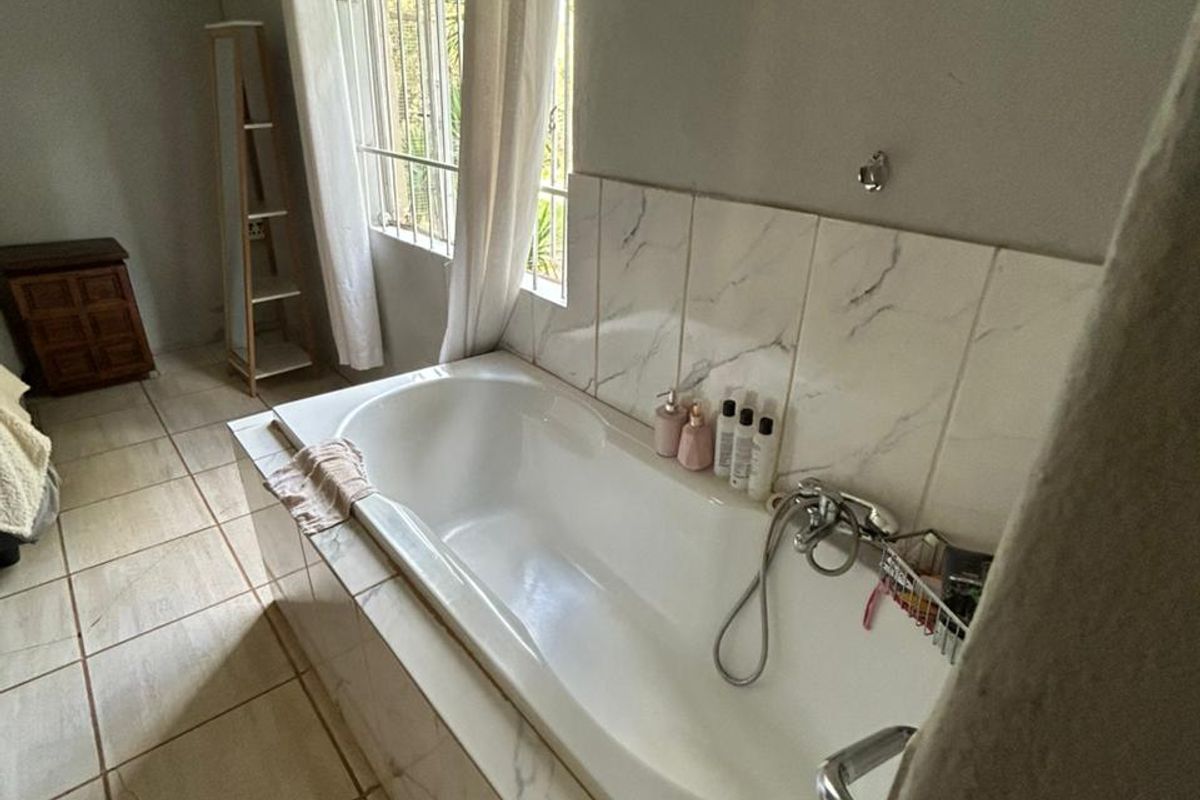 A bathroom featuring a built-in bathtub with marble-look tile surround, tiled flooring, and a window with security bars.