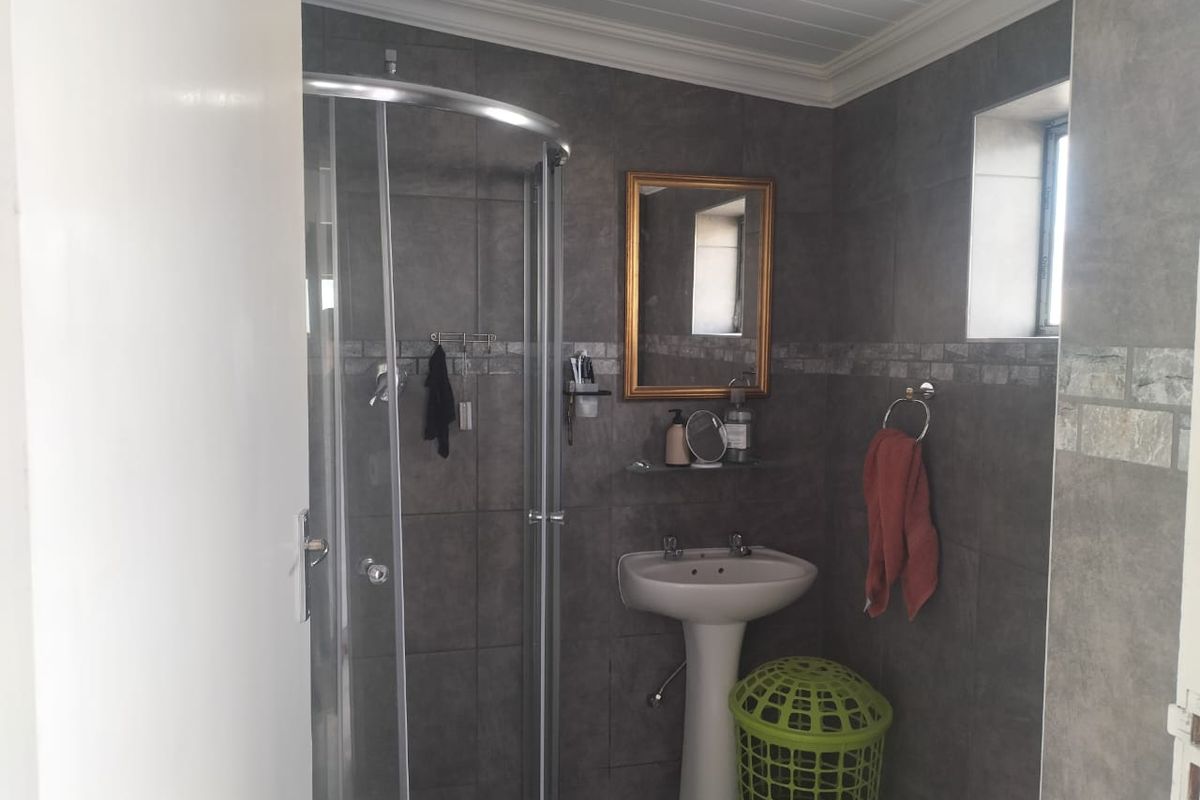 A compact bathroom featuring grey wall tiles, a pedestal basin, and a corner glass shower enclosure with a white tongue-and-groove ceiling.