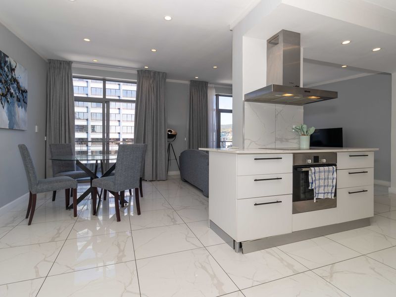 Modern open-plan living area featuring sleek white kitchen cabinetry, integrated appliances, marble-look tiled flooring, and large sliding doors leading to a balcony.