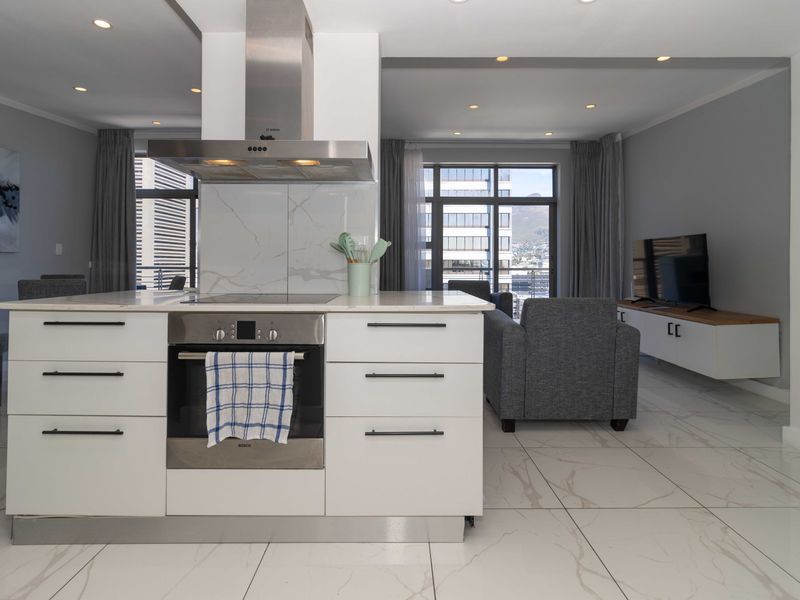 Modern open-plan living area featuring a sleek kitchen island with built-in oven, cooktop, and extractor, complemented by polished marble-look floor tiles and recessed lighting.