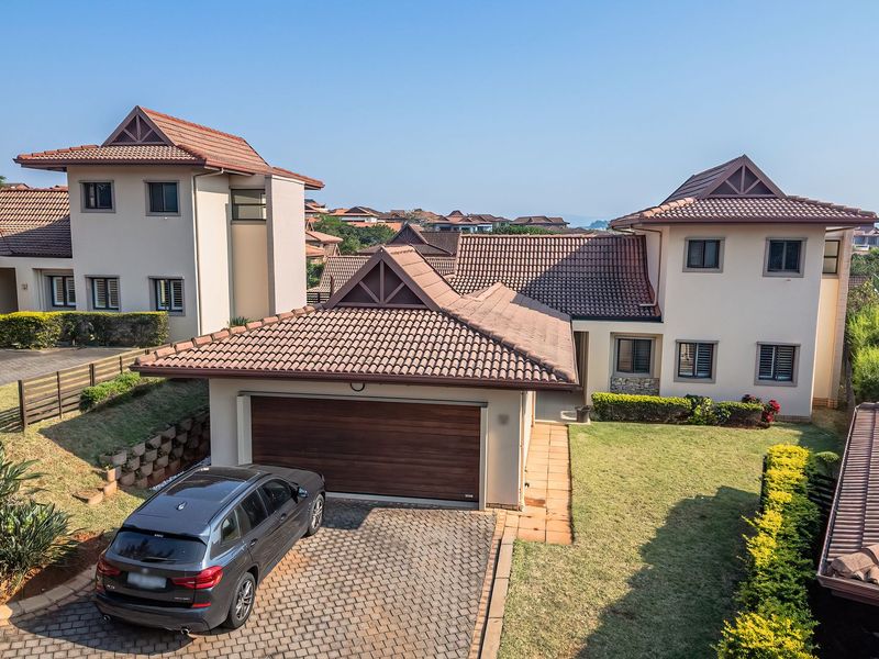 Aerial view of a residential property featuring a tiled roof, paved driveway, attached garage, and landscaped garden areas.