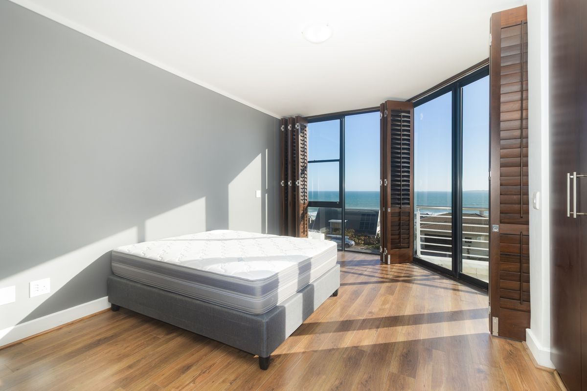 Bright bedroom featuring timber flooring, built-in cupboards, and floor-to-ceiling glass doors with timber shutters opening to a scenic ocean view.