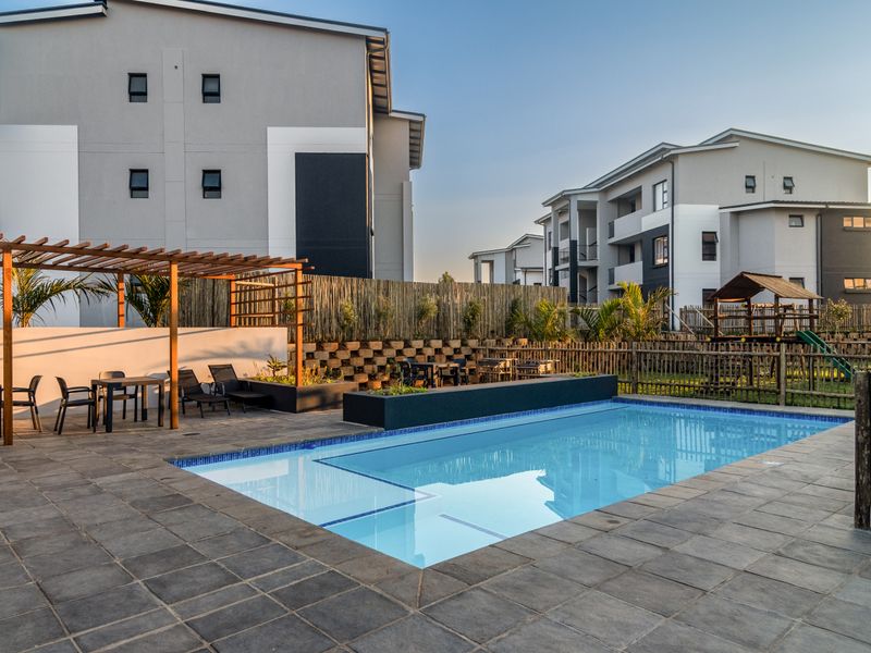 A well-maintained communal swimming pool area featuring a tiled deck, timber pergola, and landscaped retaining walls within a modern apartment complex.