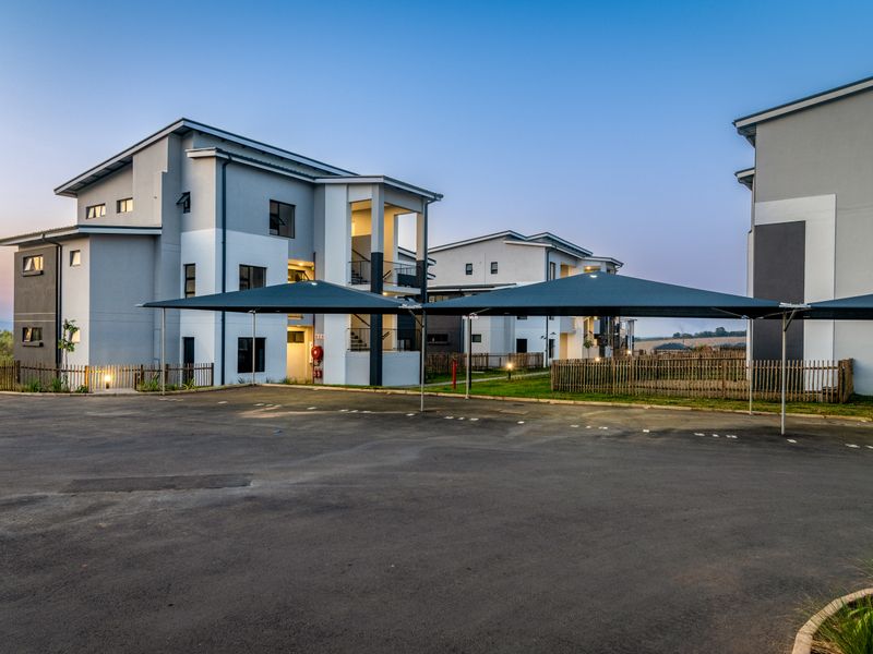 Exterior view of modern multi-story apartment buildings featuring paved parking areas with shade cloth carports and perimeter timber fencing.