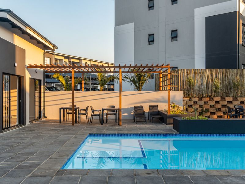 A well-maintained outdoor pool area featuring stone paving, a wooden pergola, and built-in planter boxes, set within a modern apartment complex.