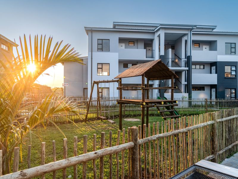 Exterior view of a modern apartment complex featuring a landscaped garden area with a wooden children's play structure and picket fencing.