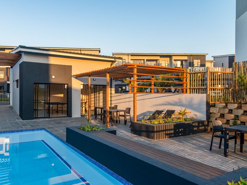 A well-maintained outdoor pool area featuring a timber pergola, paved patio, and raised planter boxes, adjacent to a modern apartment building.