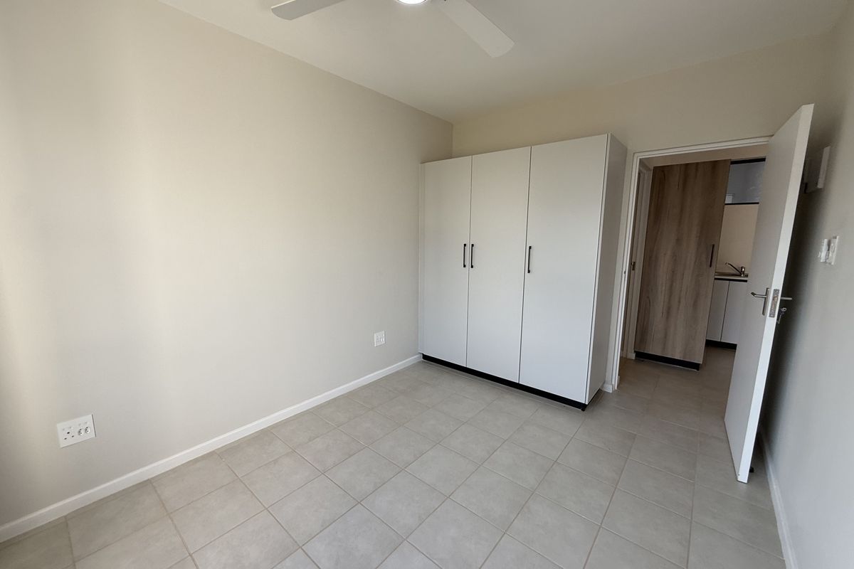 A clean, empty bedroom featuring neutral tiled flooring, a built-in white wardrobe, and a ceiling fan, with a view into an adjacent room.