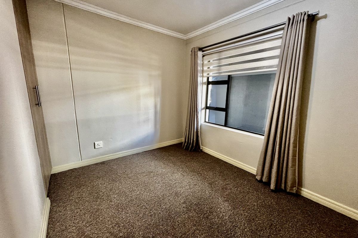 A clean, empty bedroom featuring neutral walls, carpet flooring, a built-in wardrobe, and a window with fitted blinds.