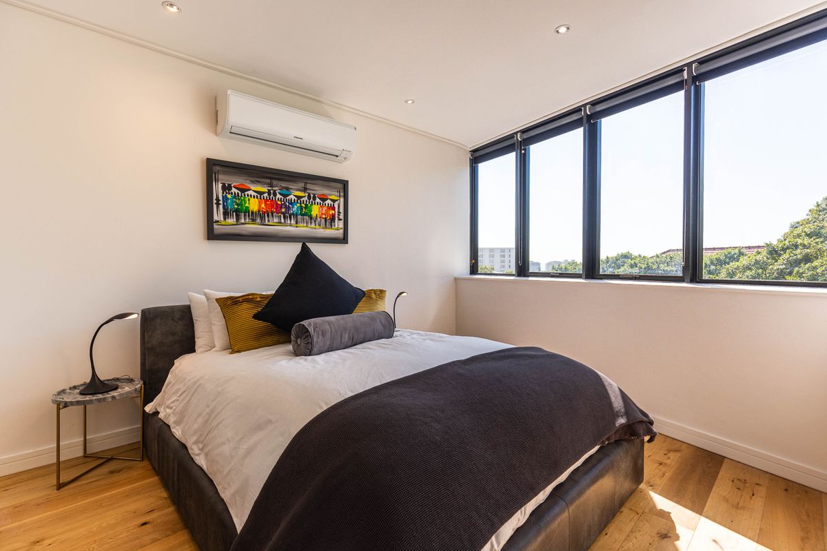 A bright and modern bedroom featuring timber flooring, large windows with natural light, and a wall-mounted air conditioning unit.