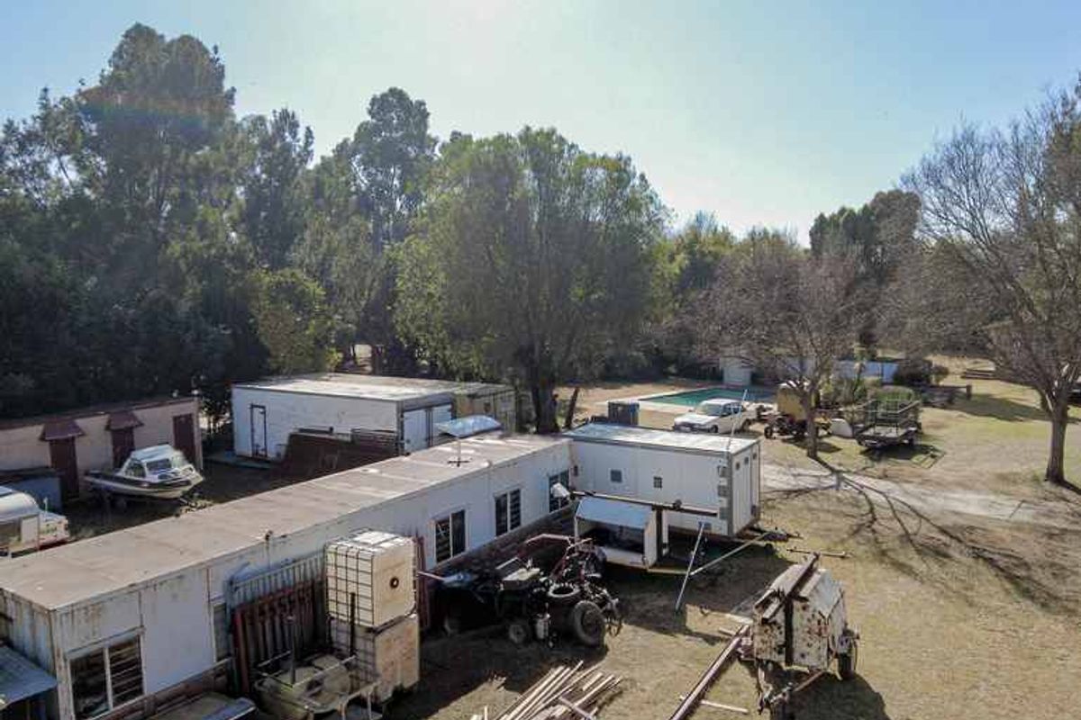 Aerial view of a small holding property featuring multiple modular structures, a swimming pool, and extensive open land with various storage and utility areas.