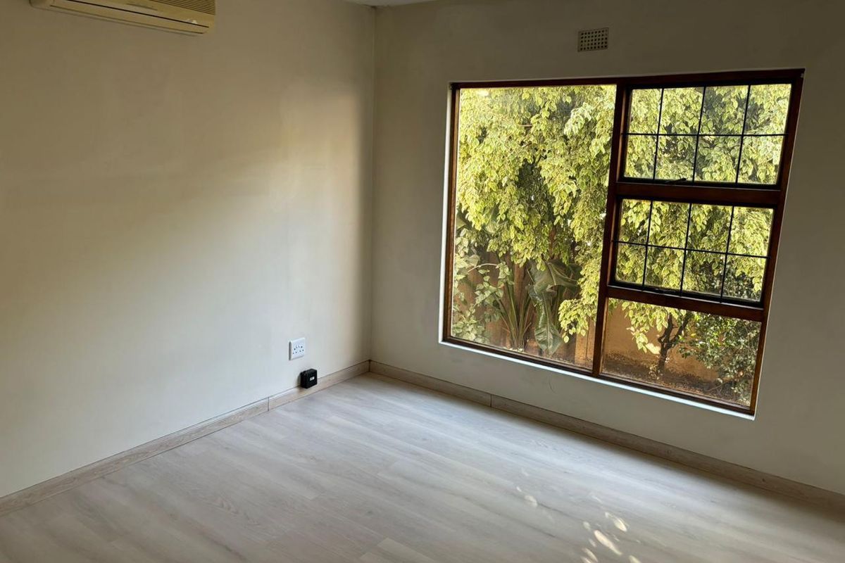 A bright, empty bedroom featuring light wood-look flooring, a large window with garden views, and a wall-mounted air conditioning unit.