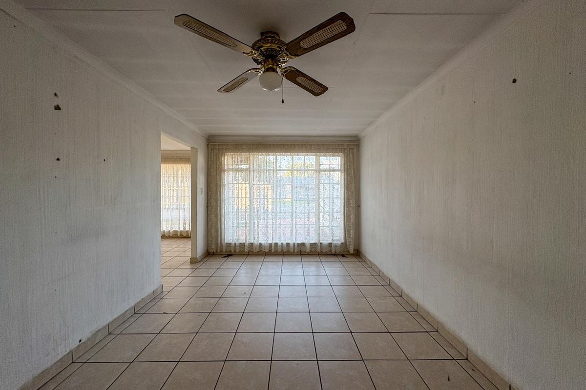 An empty, dated living area featuring tiled flooring, a ceiling fan, and large windows with curtains, offering a blank canvas for renovation.