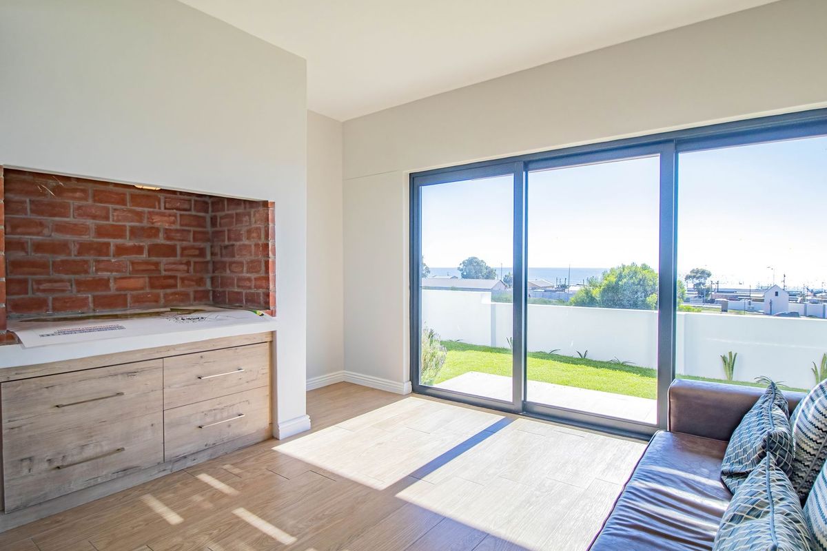 A bright living area featuring a built-in brick fireplace, wood-look flooring, and large sliding glass doors opening to a garden with coastal views.