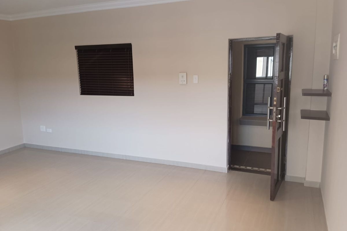 A bright, empty living area featuring neutral wall tones, tiled flooring, a dark wood window blind, and a doorway leading to an adjacent space.