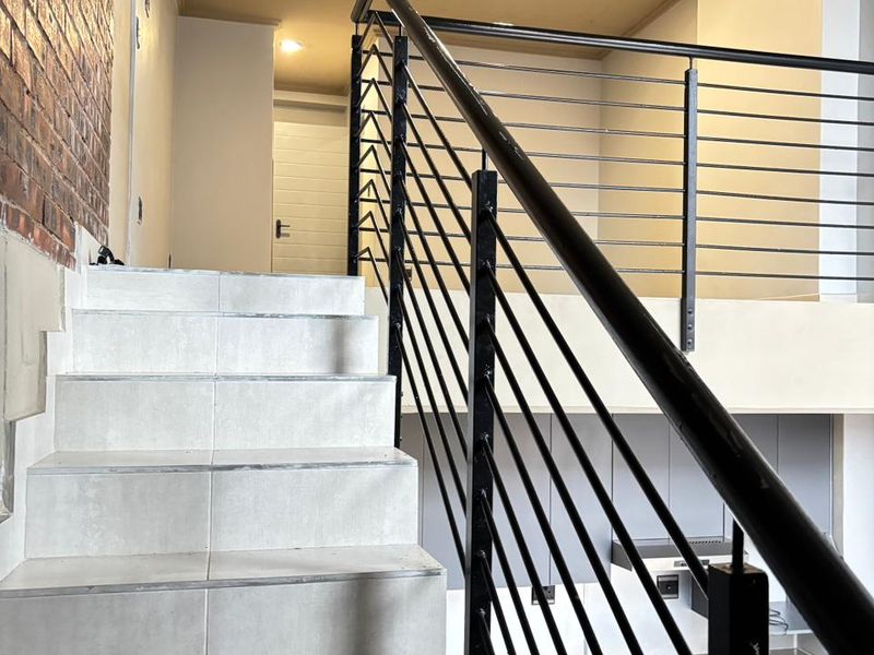 A modern apartment staircase featuring tiled steps, a sleek black metal balustrade, and an exposed brick feature wall leading to an upper mezzanine level.