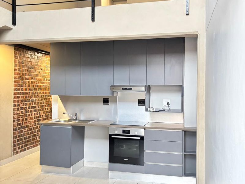 Modern kitchen featuring grey cabinetry, built-in oven, hob, and extractor fan, set against a feature brick wall with an open-plan mezzanine level above.