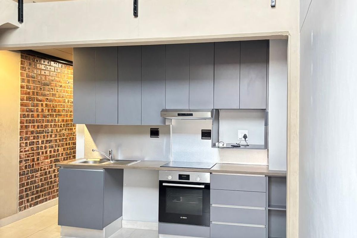 Modern kitchen featuring grey cabinetry, built-in oven, hob, and extractor fan, set against a feature brick wall with an open-plan mezzanine level above.