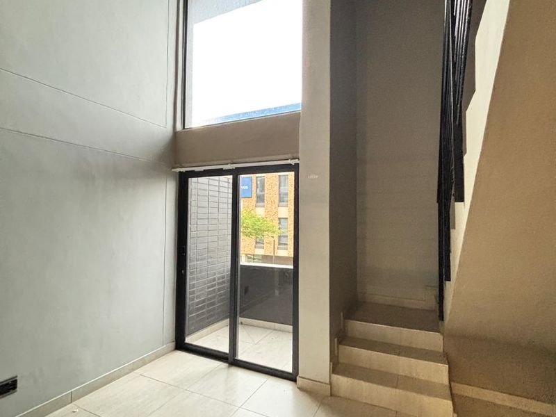 Double-volume living area featuring large windows, tiled flooring, and an internal staircase leading to the upper level, with access to a private balcony.
