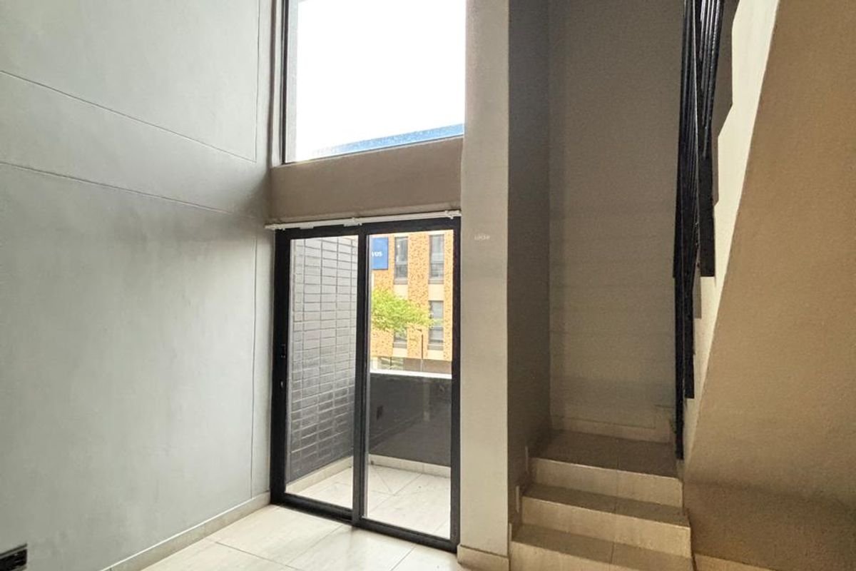Double-volume living area featuring large windows, tiled flooring, and an internal staircase leading to the upper level, with access to a private balcony.