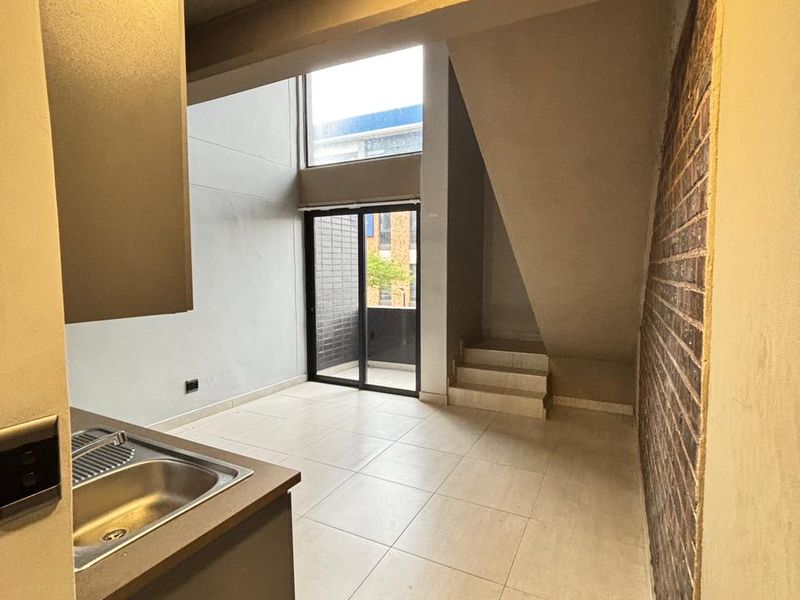 Open plan living area featuring a kitchen corner, tiled flooring, exposed brick feature wall, and a staircase leading to an upper level.