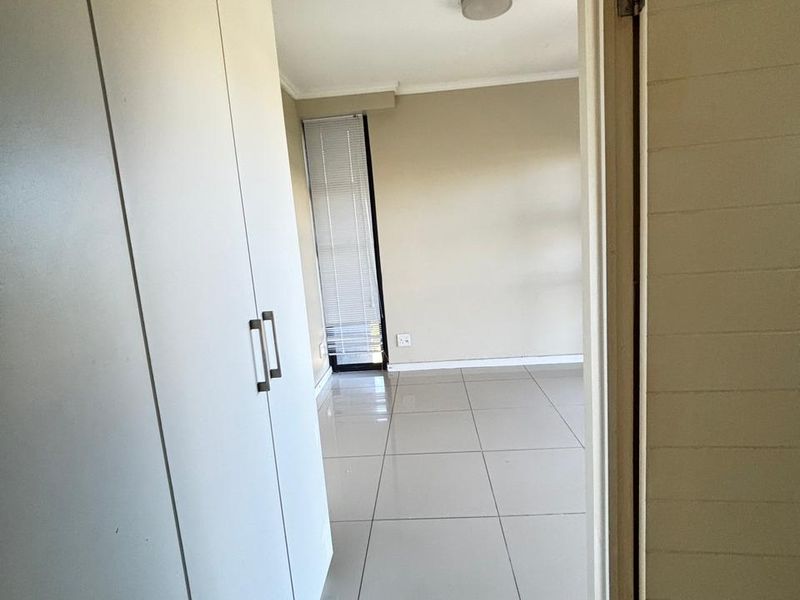 A bright, empty bedroom featuring neutral tiled flooring, built-in cupboards, and a window with fitted blinds.