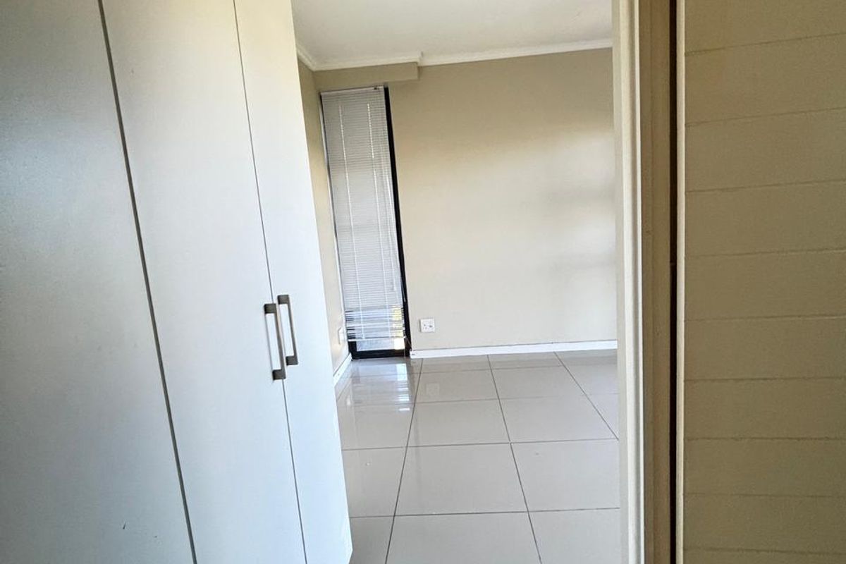 A bright, empty bedroom featuring neutral tiled flooring, built-in cupboards, and a window with fitted blinds.