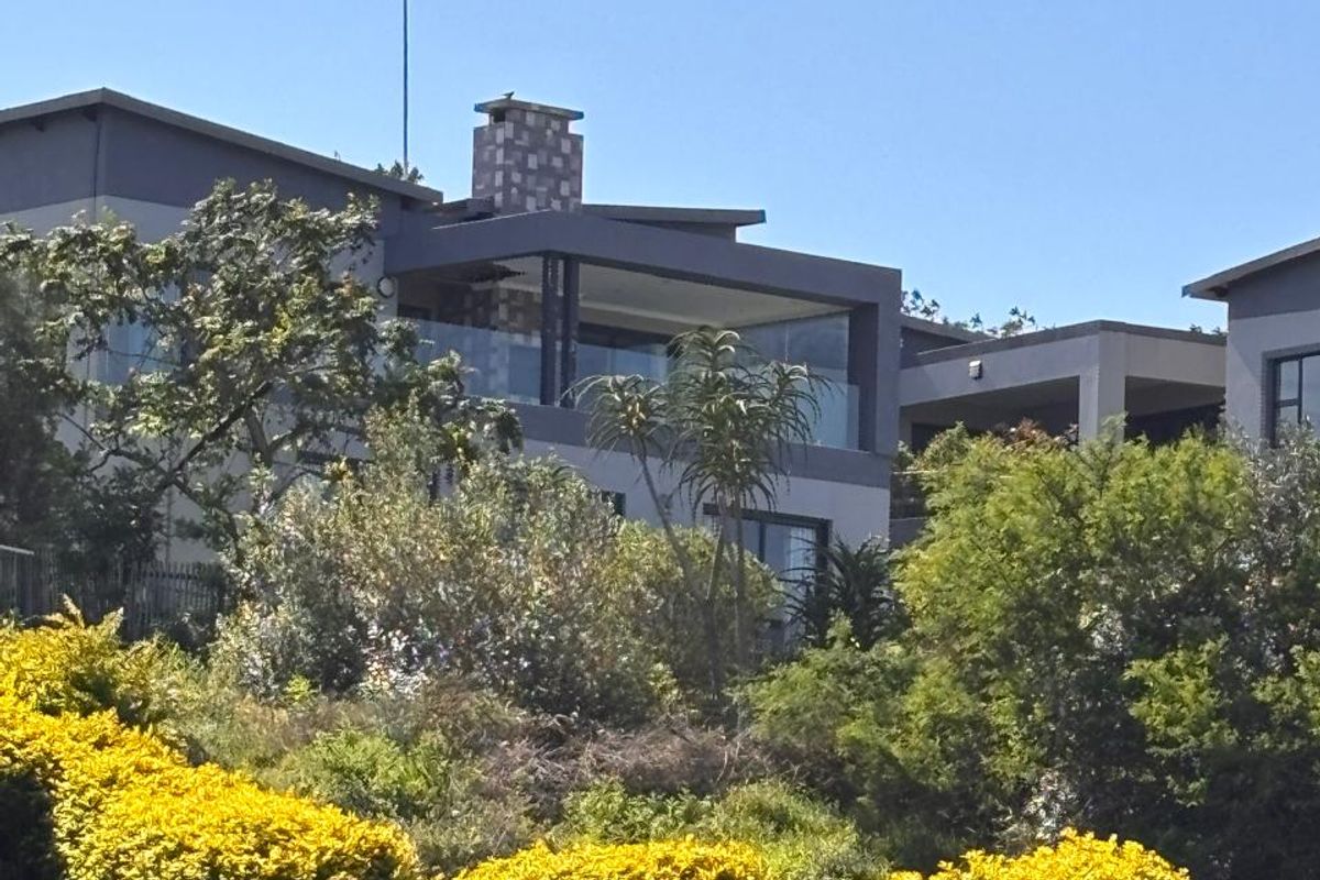 A view of the rear elevation of a modern multi-story house featuring a covered balcony, glass balustrades, and a chimney, set against lush landscaping.