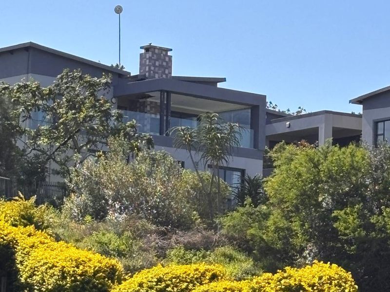 A view of the rear elevation of a modern multi-story house featuring a covered balcony, glass balustrades, and a chimney, set against lush landscaping.