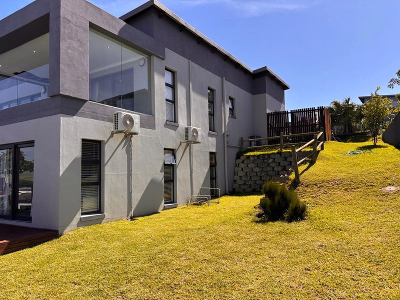 Rear view of a modern two-story house featuring a landscaped garden, retaining wall, timber decking, and glass balcony balustrade.