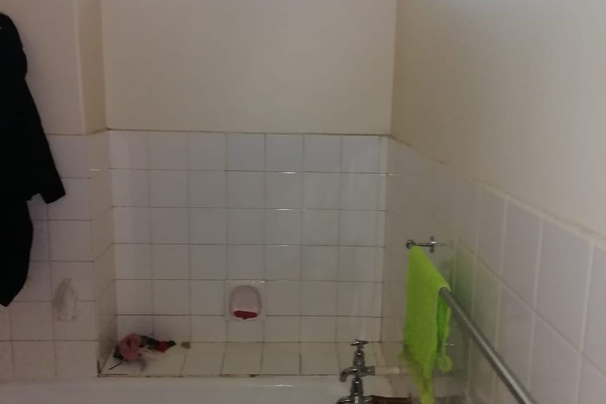A functional bathroom featuring a standard bathtub with wall-mounted taps, white tiled splashbacks, and a high-level window with security bars.