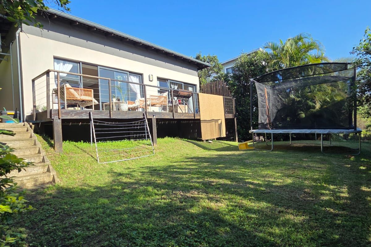Rear view of the property featuring a raised timber deck with sliding glass doors, concrete stairs, and a spacious backyard lawn.
