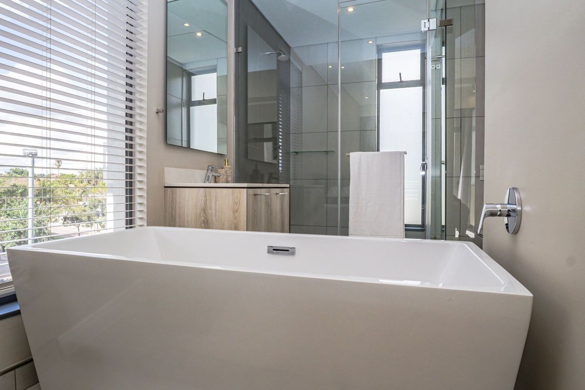 Modern bathroom featuring a freestanding bathtub, glass-enclosed shower, vanity with mirror, and large window with blinds providing natural light.