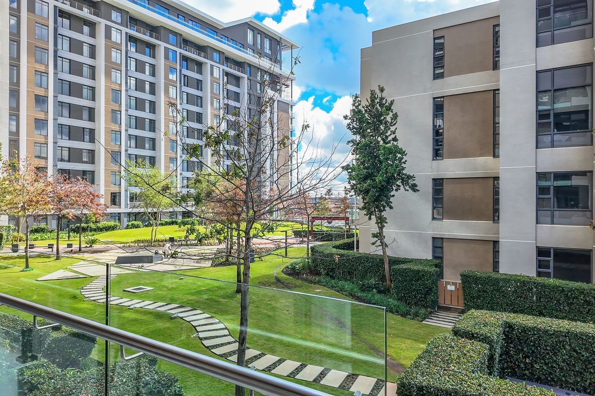 View from a balcony overlooking a well-maintained communal garden area with paved pathways, manicured hedges, and modern apartment building facades.