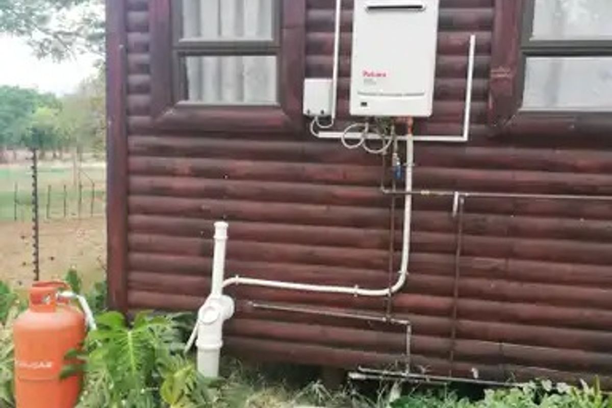 Exterior view of a timber log cabin featuring a wall-mounted gas geyser unit and associated external plumbing infrastructure.