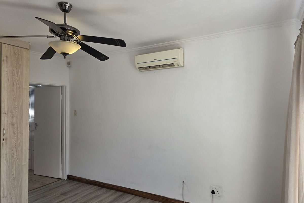 A bedroom featuring wood-look laminate flooring, a ceiling fan, and a wall-mounted air conditioning unit, with an adjacent doorway leading to another room.
