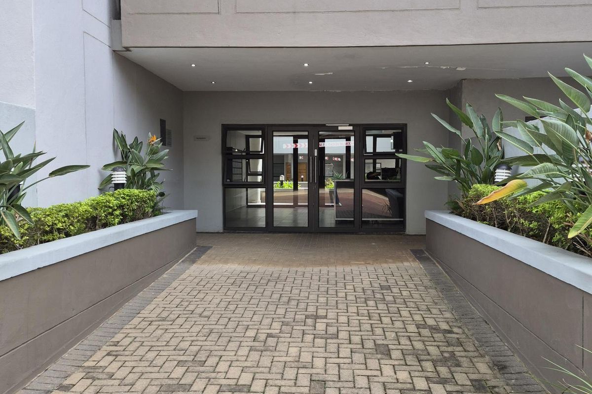 A paved walkway leads to the main entrance of the apartment building, featuring structured garden beds and a modern glass-panelled door system.
