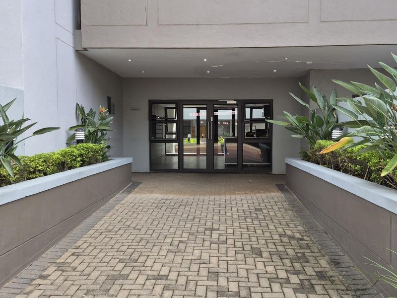 A paved walkway leads to the main entrance of the apartment building, featuring structured garden beds and a modern glass-panelled door system.