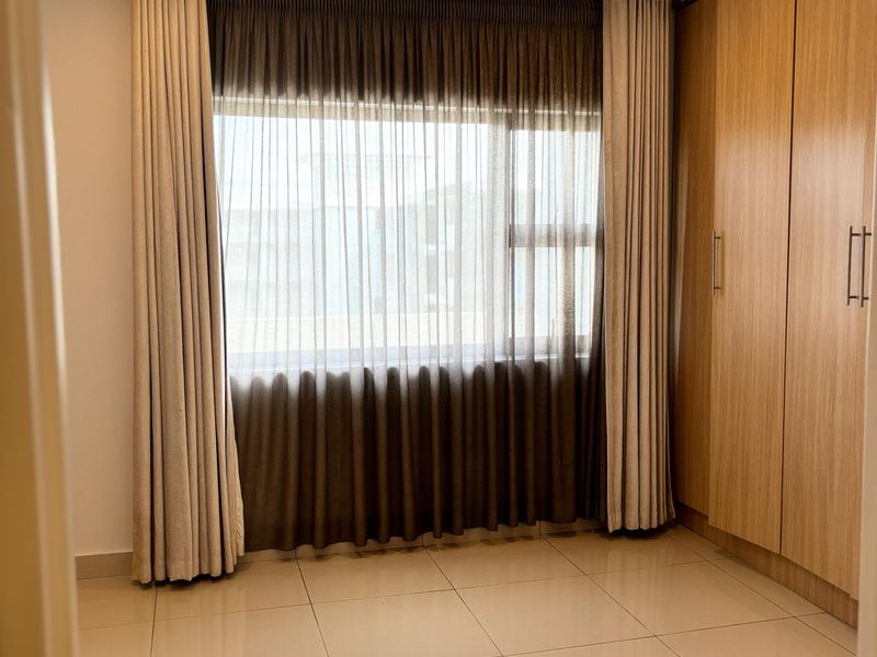 A bright bedroom featuring large format floor tiles, built-in wooden cupboards, and floor-to-ceiling window treatments.