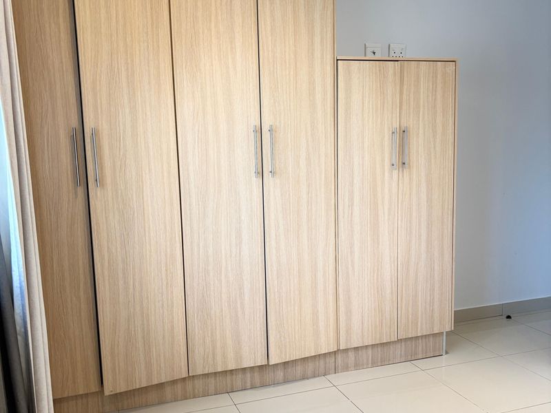 A bedroom featuring light wood-grain built-in cupboards with modern metal handles and neutral tiled flooring.