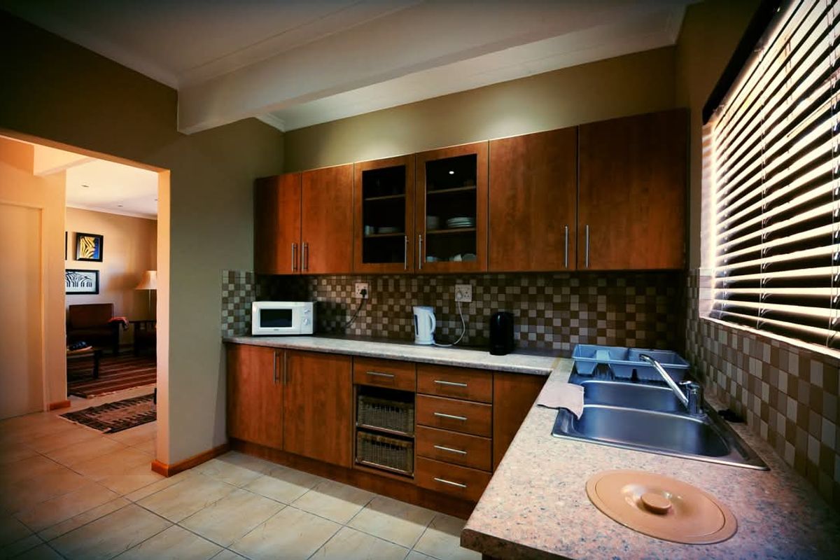 A functional kitchen featuring wooden cabinetry, tiled backsplash, stone-look countertops, and a double-basin stainless steel sink with a view into the adjacent living area.