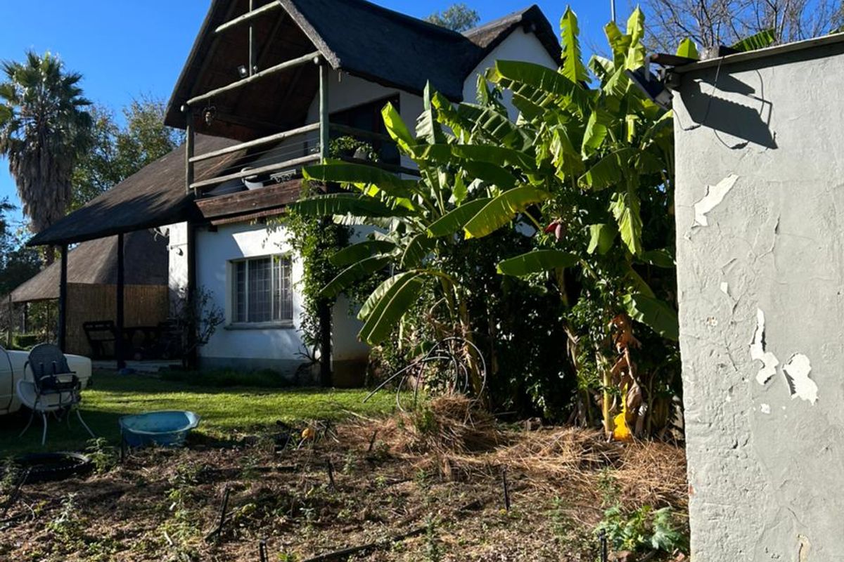 Rear view of a two-story farmhouse featuring a thatched roof, a wooden balcony, and an adjacent garden area with an outbuilding.