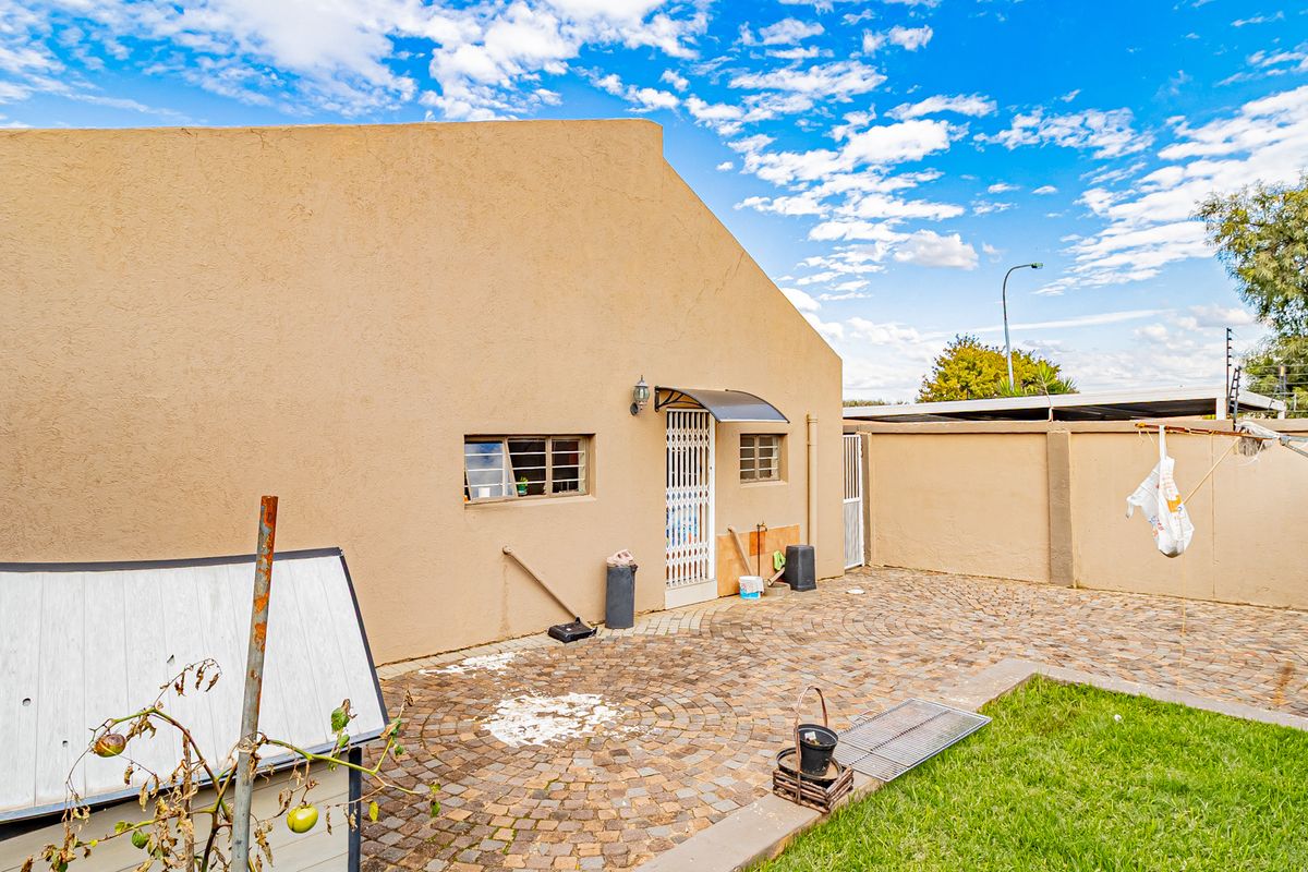 A rear exterior view of the property featuring a paved courtyard, a small grass area, and a secure entrance door with a protective awning.