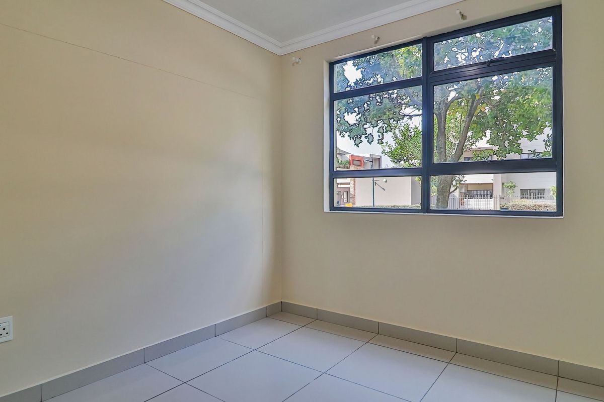 A bright, empty bedroom featuring neutral tiled flooring, cream-colored walls, and a large window with dark frames overlooking the exterior.
