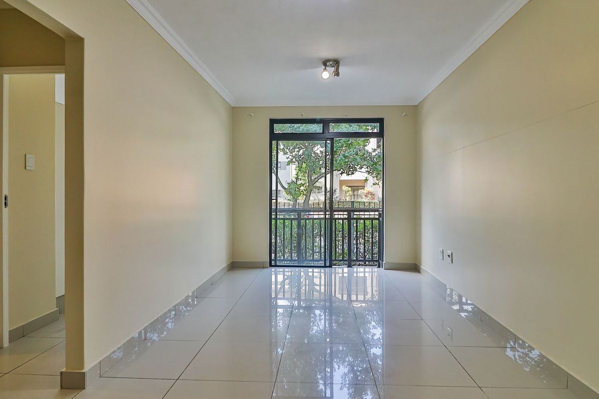 A bright, empty living area featuring polished floor tiles, neutral walls, and a sliding glass door leading to a balcony with garden views.